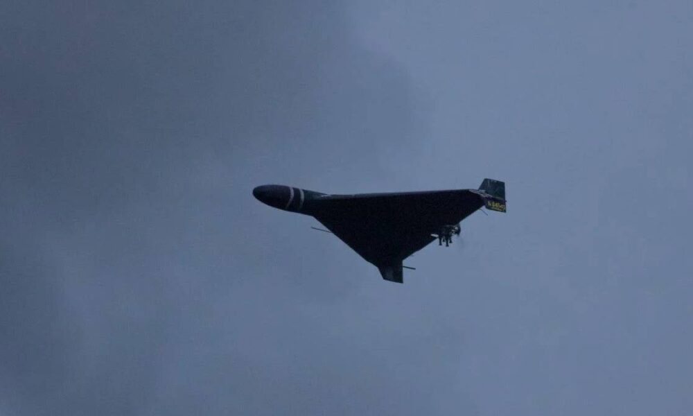 Dark triangular stealth bomber flying wing silhouette against a cloudy blue sky, with landing gear extended cradled beneath.