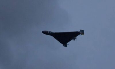 Dark triangular stealth bomber flying wing silhouette against a cloudy blue sky, with landing gear extended cradled beneath.