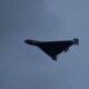 Dark triangular stealth bomber flying wing silhouette against a cloudy blue sky, with landing gear extended cradled beneath.