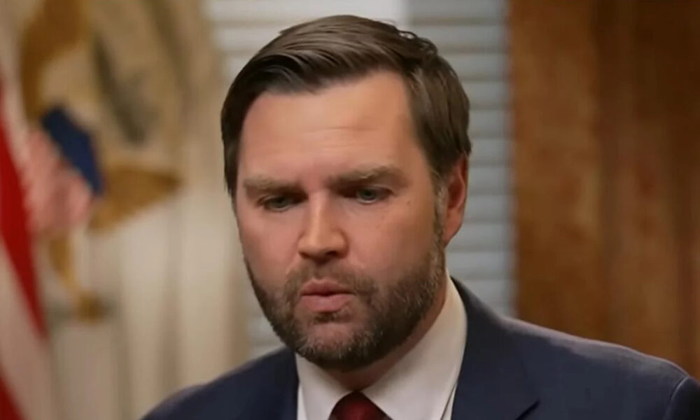 Man in a navy suit with a red tie speaking, seen from the chest up in a hallway setting