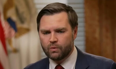 Man in a navy suit with a red tie speaking, seen from the chest up in a hallway setting