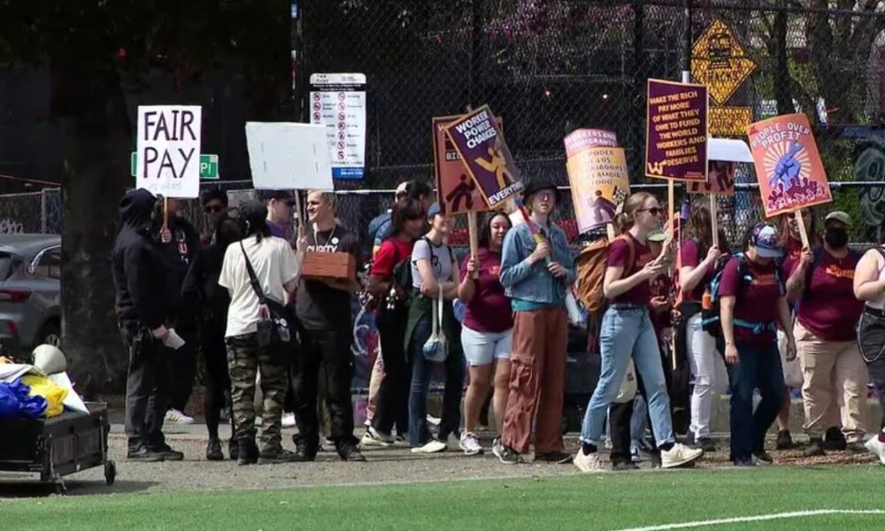 Group of protesters walking along a fence line, holding signs like 'FAIR PAY' and other slogans in a park setting.