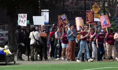 Group of protesters walking along a fence line, holding signs like 'FAIR PAY' and other slogans in a park setting.