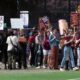 Group of protesters walking along a fence line, holding signs like 'FAIR PAY' and other slogans in a park setting.