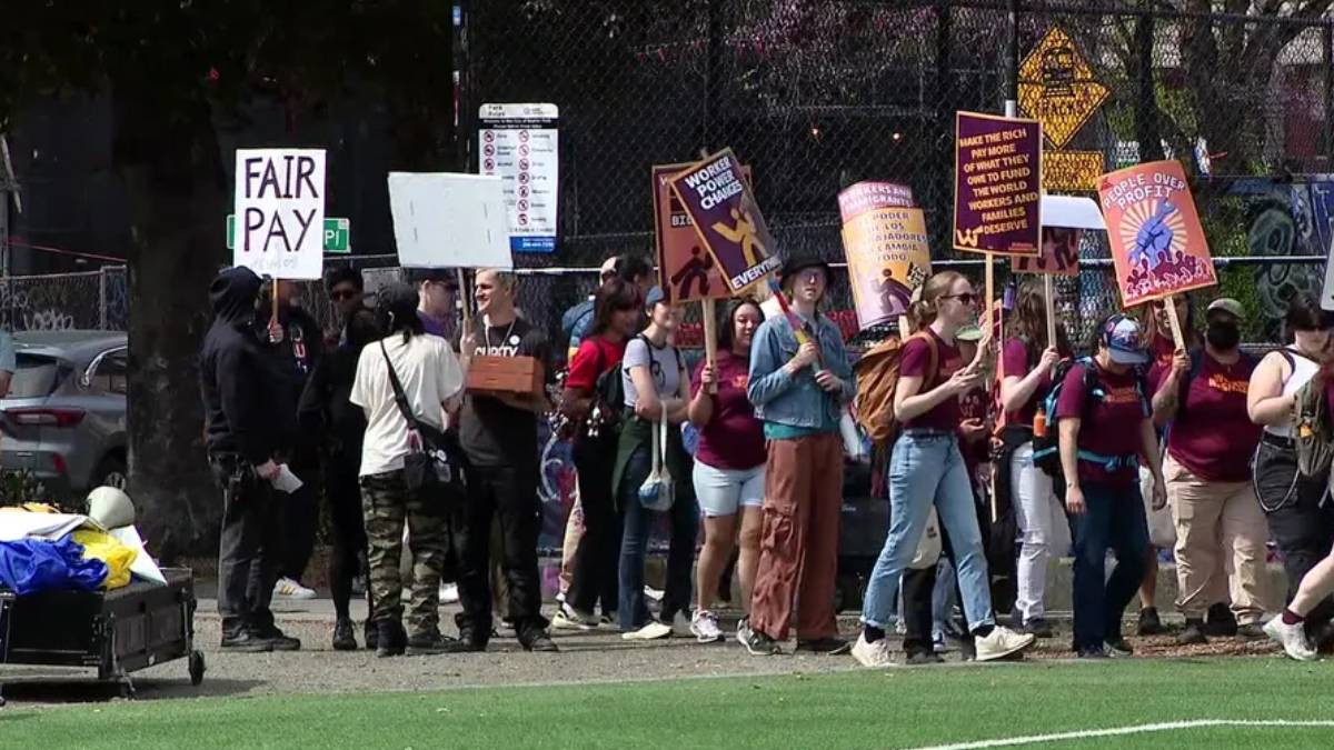Group of protesters walking along a fence line, holding signs like 'FAIR PAY' and other slogans in a park setting.