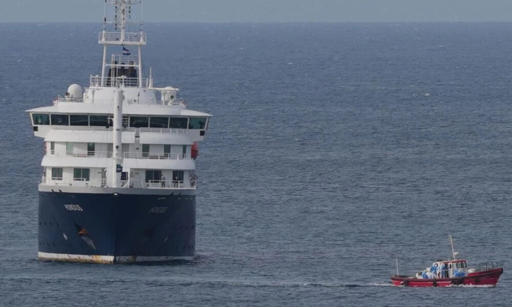 Large white and blue passenger ferry on calm open water, with a small red boat nearby for scale and support.