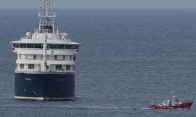 Large white and blue passenger ferry on calm open water, with a small red boat nearby for scale and support.
