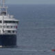 Large white and blue passenger ferry on calm open water, with a small red boat nearby for scale and support.