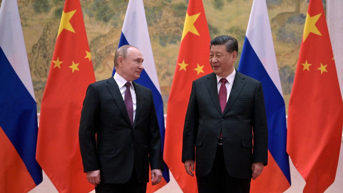 Two male political leaders in dark suits stand facing each other in front of Russia and China flags.