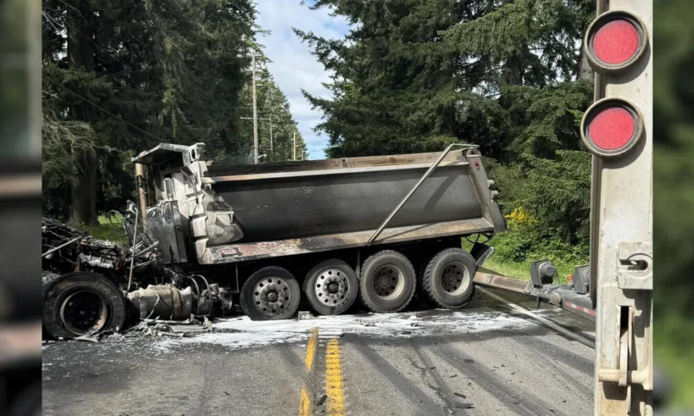 Dump truck crashed on a two-lane road at a railroad crossing; cab crushed and debris scattered, red crossing lights nearby.