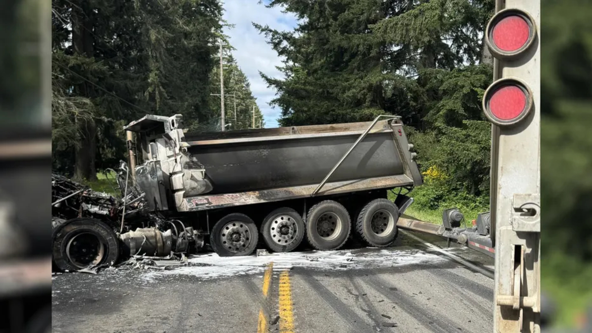 Dump truck crashed on a two-lane road at a railroad crossing; cab crushed and debris scattered, red crossing lights nearby.
