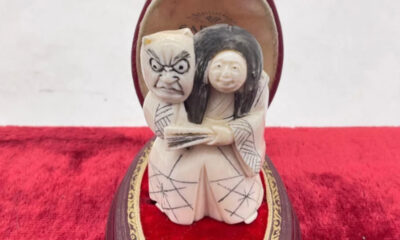 Carved ivory sculpture of a grimacing figure and a smiling woman in traditional robes, embraced on a red velvet base.