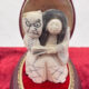 Carved ivory sculpture of a grimacing figure and a smiling woman in traditional robes, embraced on a red velvet base.
