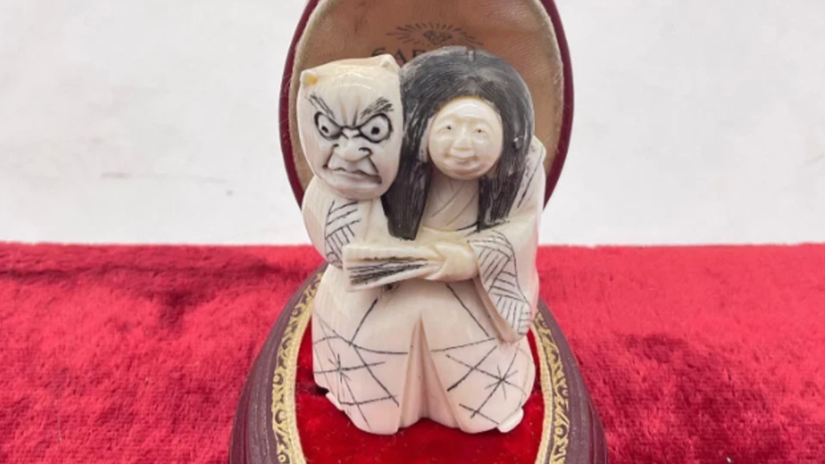 Carved ivory sculpture of a grimacing figure and a smiling woman in traditional robes, embraced on a red velvet base.