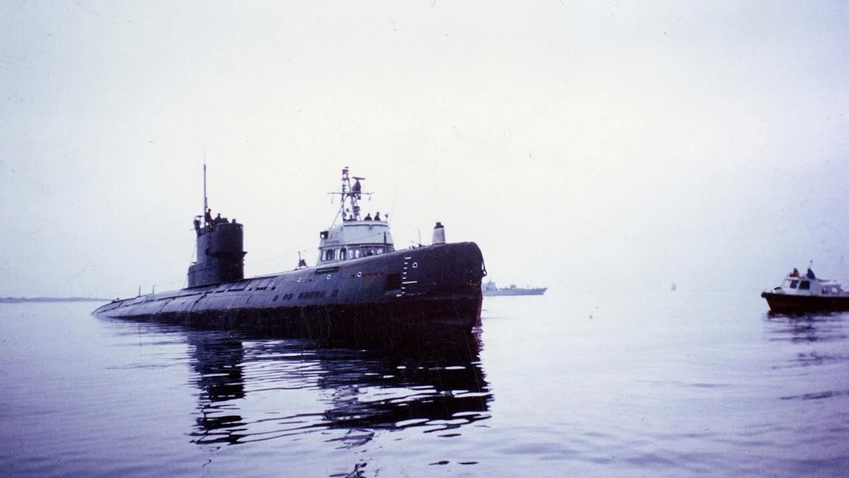 Submarine partially surfaced in calm water, with a small boat to the right and reflections on the surface.