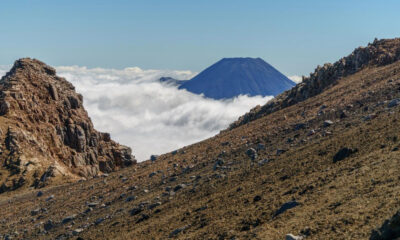 Volcanic landscape with rocky slopes and a distant cone-shaped volcano rising above a sea of clouds under a blue sky.