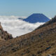 Volcanic landscape with rocky slopes and a distant cone-shaped volcano rising above a sea of clouds under a blue sky.
