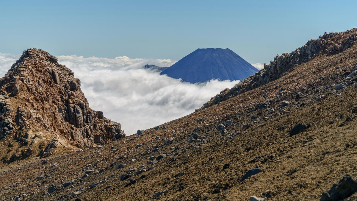 Volcanic landscape with rocky slopes and a distant cone-shaped volcano rising above a sea of clouds under a blue sky.