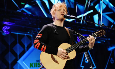 Male singer with blonde hair performing live on stage, playing an acoustic guitar. Singer with a black shirt and red stripe sleeves on stage amid blue lights, playing guitar into a mic. A musician strums an acoustic guitar into a microphone during a concert. Live stage performance: vocalist singing into a mic while playing an acoustic guitar. Close-up of a guitarist singing into a microphone on a brightly lit stage.