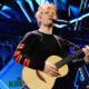 Male singer with blonde hair performing live on stage, playing an acoustic guitar. Singer with a black shirt and red stripe sleeves on stage amid blue lights, playing guitar into a mic. A musician strums an acoustic guitar into a microphone during a concert. Live stage performance: vocalist singing into a mic while playing an acoustic guitar. Close-up of a guitarist singing into a microphone on a brightly lit stage.
