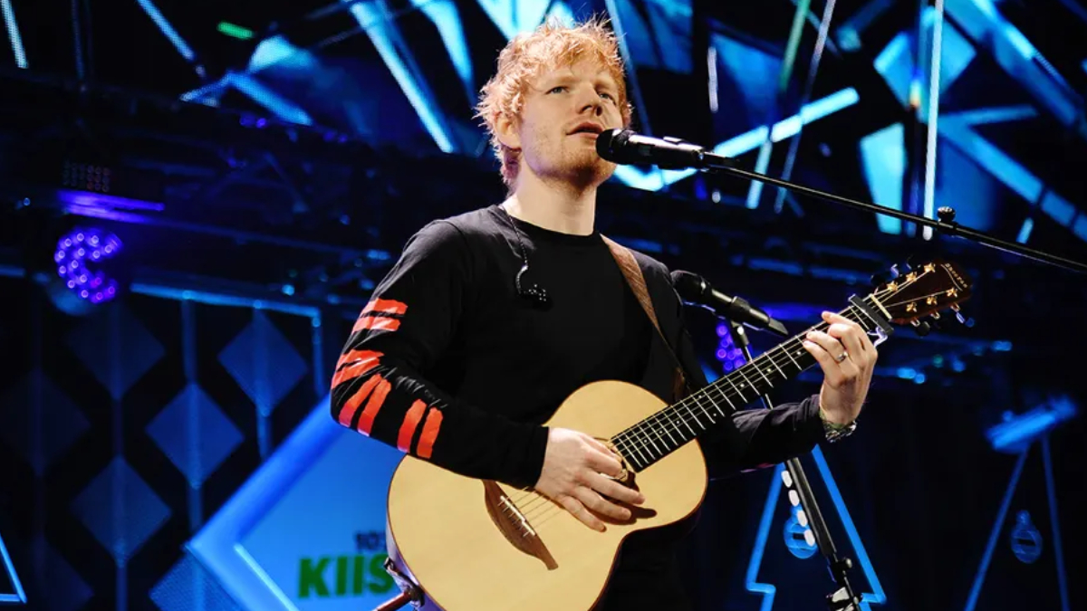 Male singer with blonde hair performing live on stage, playing an acoustic guitar. Singer with a black shirt and red stripe sleeves on stage amid blue lights, playing guitar into a mic. A musician strums an acoustic guitar into a microphone during a concert. Live stage performance: vocalist singing into a mic while playing an acoustic guitar. Close-up of a guitarist singing into a microphone on a brightly lit stage.