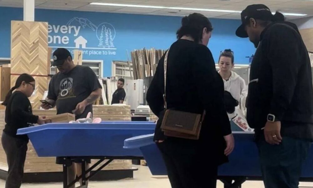 Shoppers gather around a blue display table in a home improvement store, inspecting items as a staff member assists nearby.
