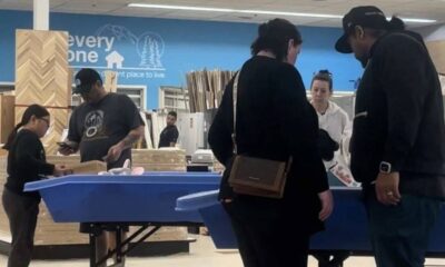 Shoppers gather around a blue display table in a home improvement store, inspecting items as a staff member assists nearby.