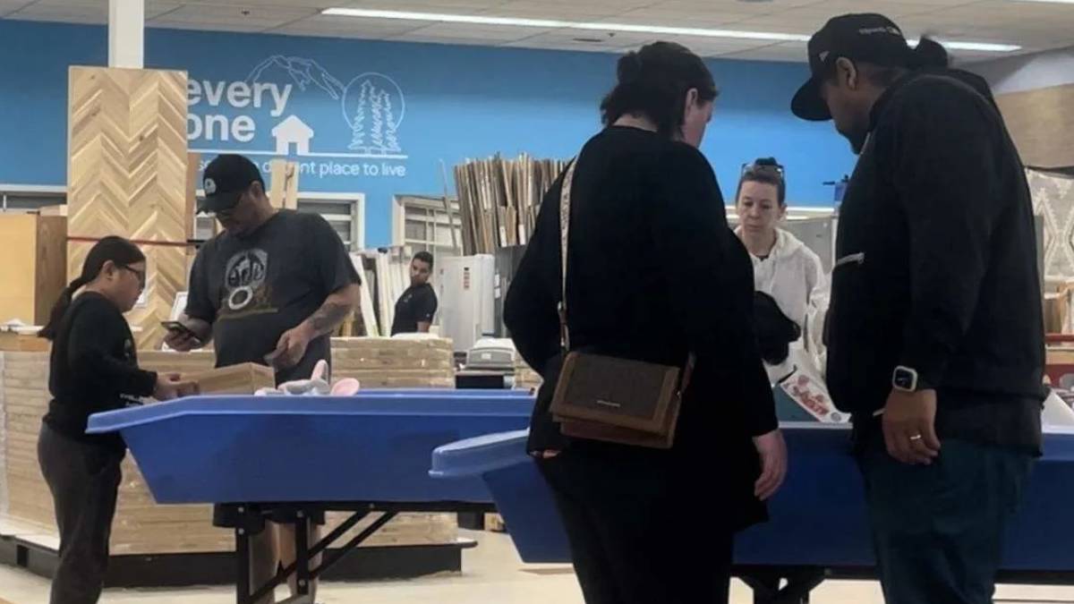 Shoppers gather around a blue display table in a home improvement store, inspecting items as a staff member assists nearby.
