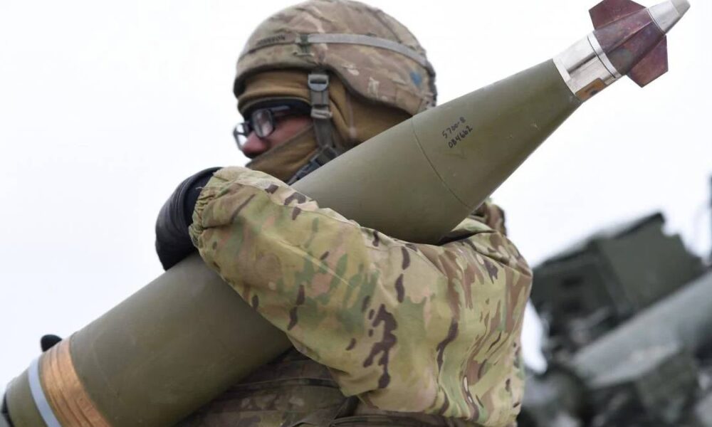 Soldier in camouflage gear and helmet holds a large green missile over his shoulder, outdoors with military equipment in the background.