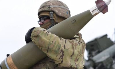 Soldier in camouflage gear and helmet holds a large green missile over his shoulder, outdoors with military equipment in the background.