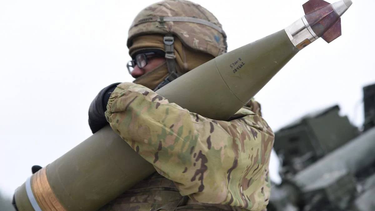 Soldier in camouflage gear and helmet holds a large green missile over his shoulder, outdoors with military equipment in the background.