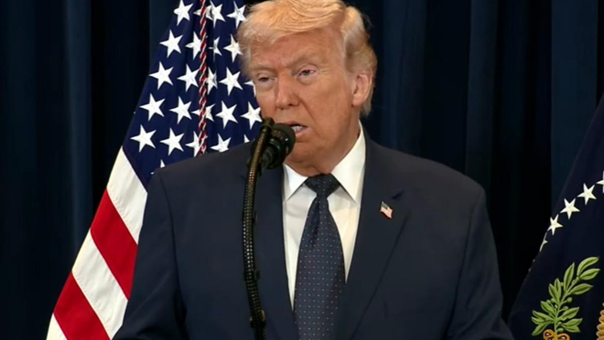 Politician in a dark suit speaks at a podium with a microphone, American flag in the background behind him
