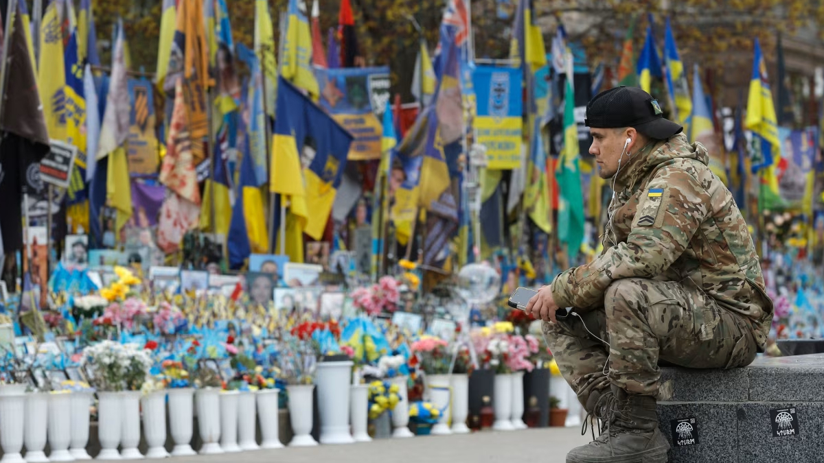 A Ukrainian soldier in camouflage sits on a stone ledge at a memorial decorated with yellow and blue flags, photos and flowers behind him.