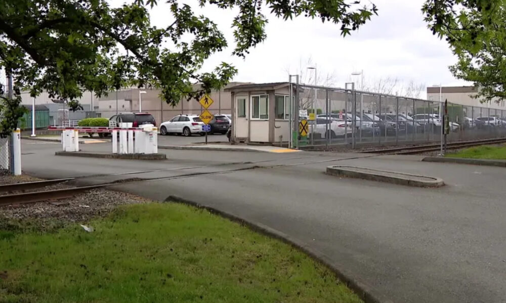 Railroad crossing entrance to a fenced industrial lot with a guard booth and parked cars nearby