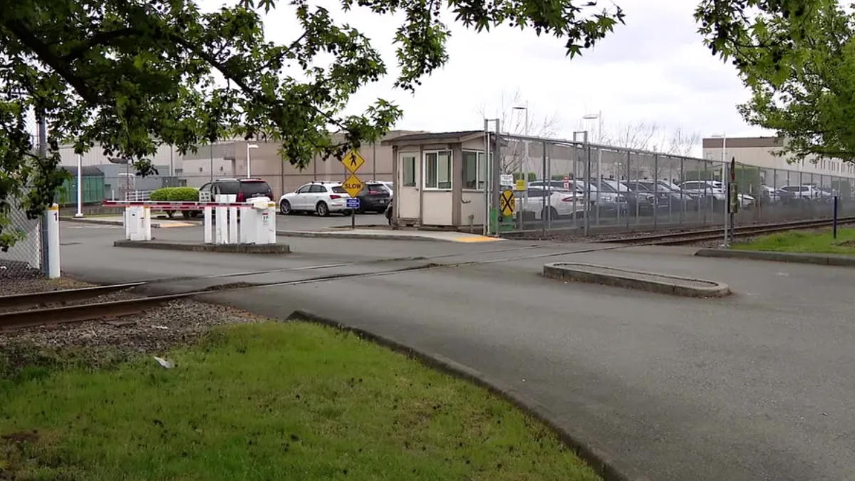 Railroad crossing entrance to a fenced industrial lot with a guard booth and parked cars nearby
