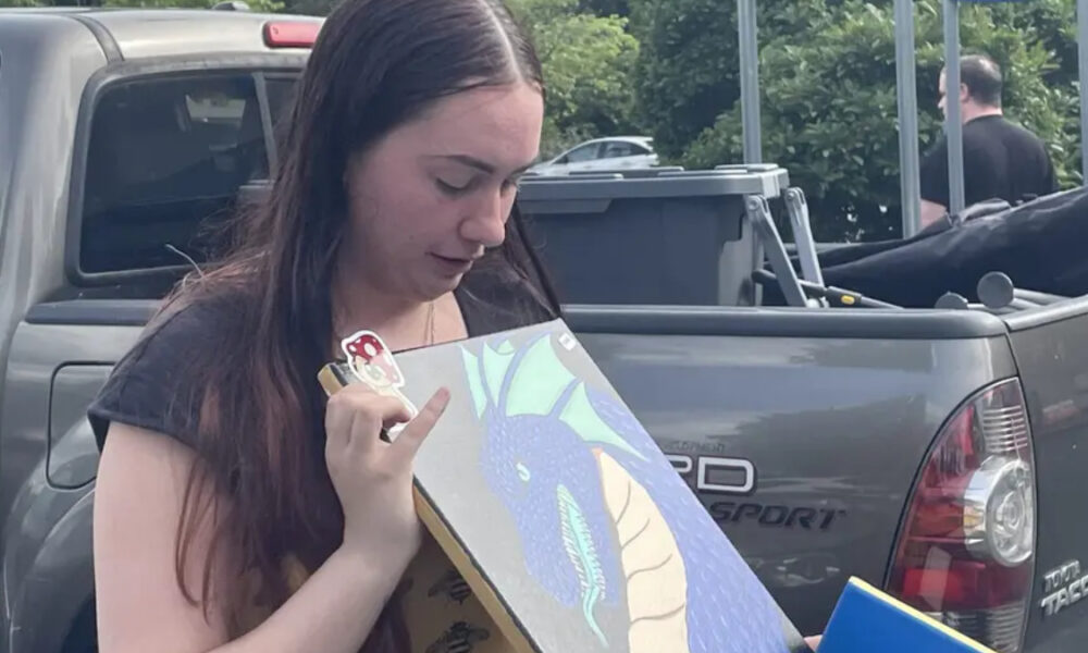 Young woman stands beside a gray pickup, looking down at a dragon illustration she is holding.