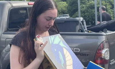 Young woman stands beside a gray pickup, looking down at a dragon illustration she is holding.