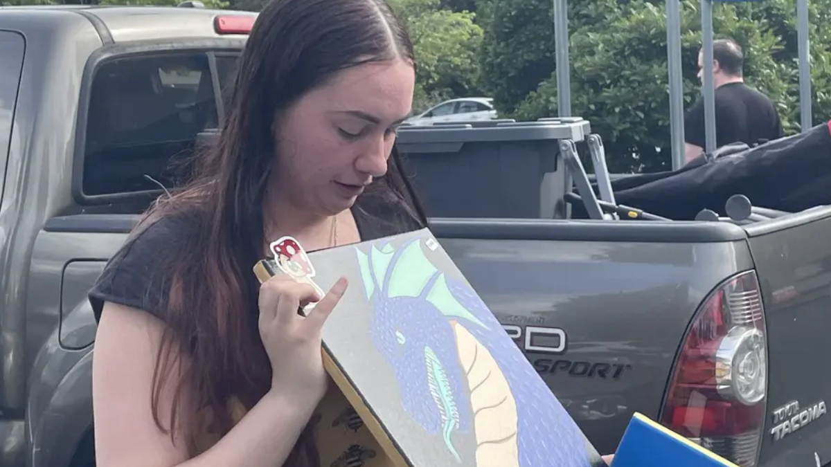 Young woman stands beside a gray pickup, looking down at a dragon illustration she is holding.