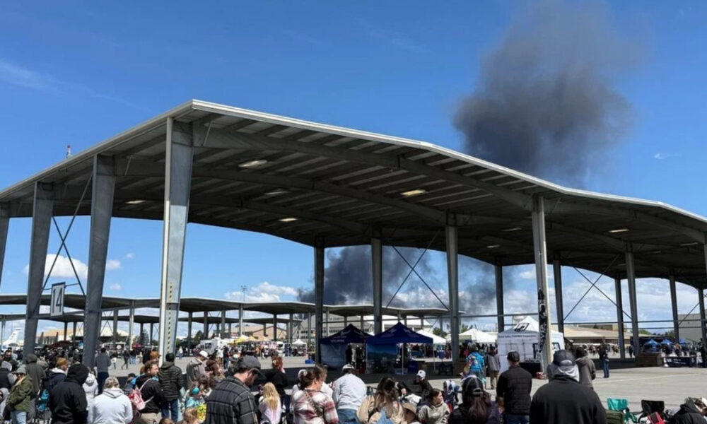 Crowd gathers at an outdoor event under a large metal pavilion, with dark smoke rising behind the structure in the distance.