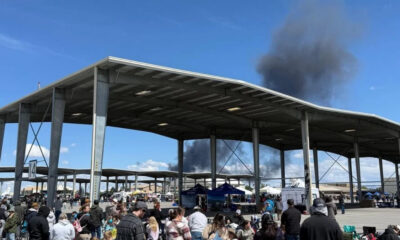 Crowd gathers at an outdoor event under a large metal pavilion, with dark smoke rising behind the structure in the distance.