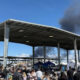 Crowd gathers at an outdoor event under a large metal pavilion, with dark smoke rising behind the structure in the distance.