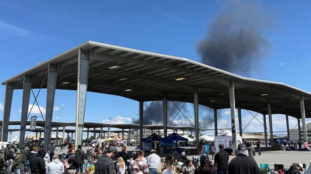 Crowd gathers at an outdoor event under a large metal pavilion, with dark smoke rising behind the structure in the distance.