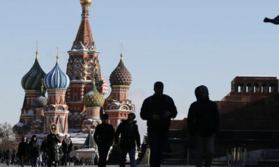 Silhouetted pedestrians walk past Saint Basil's Cathedral in Moscow with a bird flying in the clear blue sky nearby.
