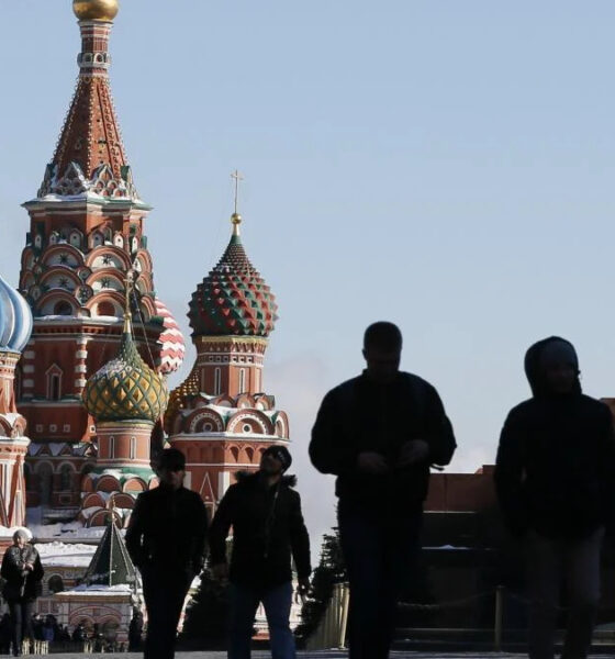 Silhouetted pedestrians walk past Saint Basil's Cathedral in Moscow with a bird flying in the clear blue sky nearby.