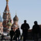 Silhouetted pedestrians walk past Saint Basil's Cathedral in Moscow with a bird flying in the clear blue sky nearby.