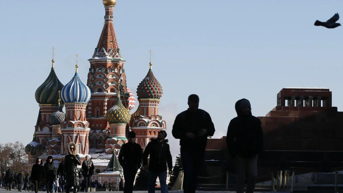 Silhouetted pedestrians walk past Saint Basil's Cathedral in Moscow with a bird flying in the clear blue sky nearby.