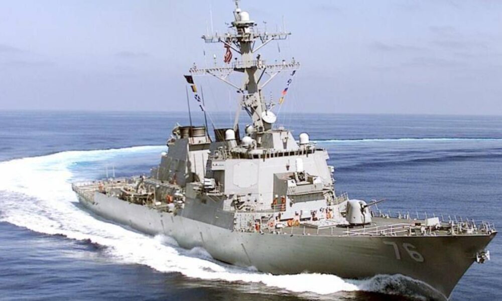 Gray military destroyer sailing at sea, bow numbers 76 visible and white wake behind it, antenna mast overhead, open water ahead.