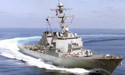 Gray military destroyer sailing at sea, bow numbers 76 visible and white wake behind it, antenna mast overhead, open water ahead.