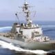 Gray military destroyer sailing at sea, bow numbers 76 visible and white wake behind it, antenna mast overhead, open water ahead.