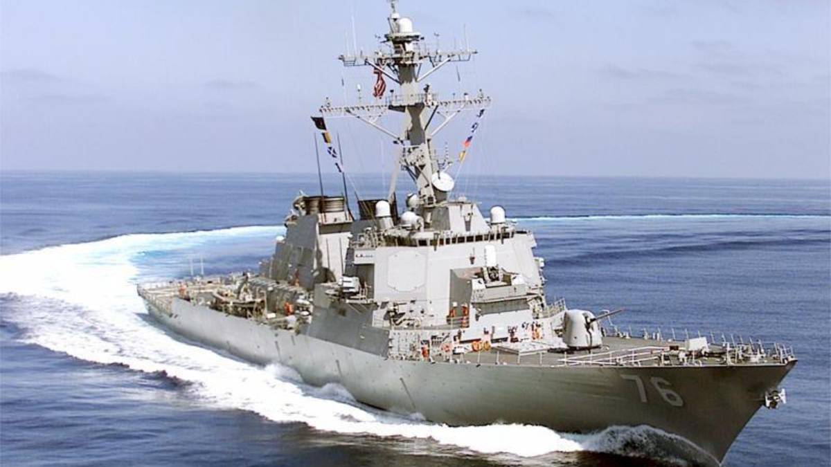 Gray military destroyer sailing at sea, bow numbers 76 visible and white wake behind it, antenna mast overhead, open water ahead.